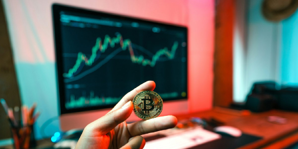 "A person holding a Bitcoin coin in front of a monitor displaying an upward trending cryptocurrency market graph.