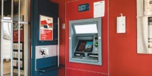 An ATM machine embedded in a red wall beside informational posters and a hand sanitizer dispenser.