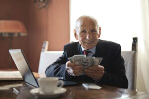 An elderly man smiling while holding a stack of cash and using a laptop, symbolizing the benefits of incorporating cryptocurrency mining into a Self-Managed Super Fund (SMSF) for retirement planning.