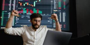 Joyful trader celebrating success in front of a laptop displaying live trading charts.