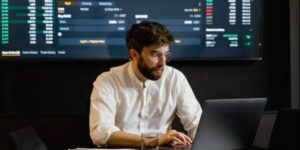 Cryptocurrency trader analyzing market data on laptop with trading dashboard in the background.