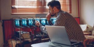 Man carefully monitoring a cryptocurrency mining rig setup in a home environment with multiple GPUs and a laptop