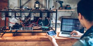 Cryptocurrency miner monitoring a custom-built GPU mining rig with a laptop and smartphone in a home workshop