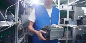Technician wearing a blue vest holding an ASIC miner inside a cryptocurrency mining facility.