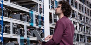 Technician monitoring cryptocurrency mining rigs with a laptop inside a bitcoin mining facility.