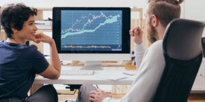 Two people analysing a Bitcoin price chart on a computer screen, discussing bitcoin cloud mining performance and returns.