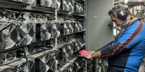 Technician inspecting ASIC miners on a rack, wearing hearing protection and gloves.
