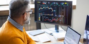 Person reading trading charts on a desktop monitor and laptop in a home office setting