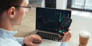 Person monitoring trading charts on a laptop and smartphone at a desk